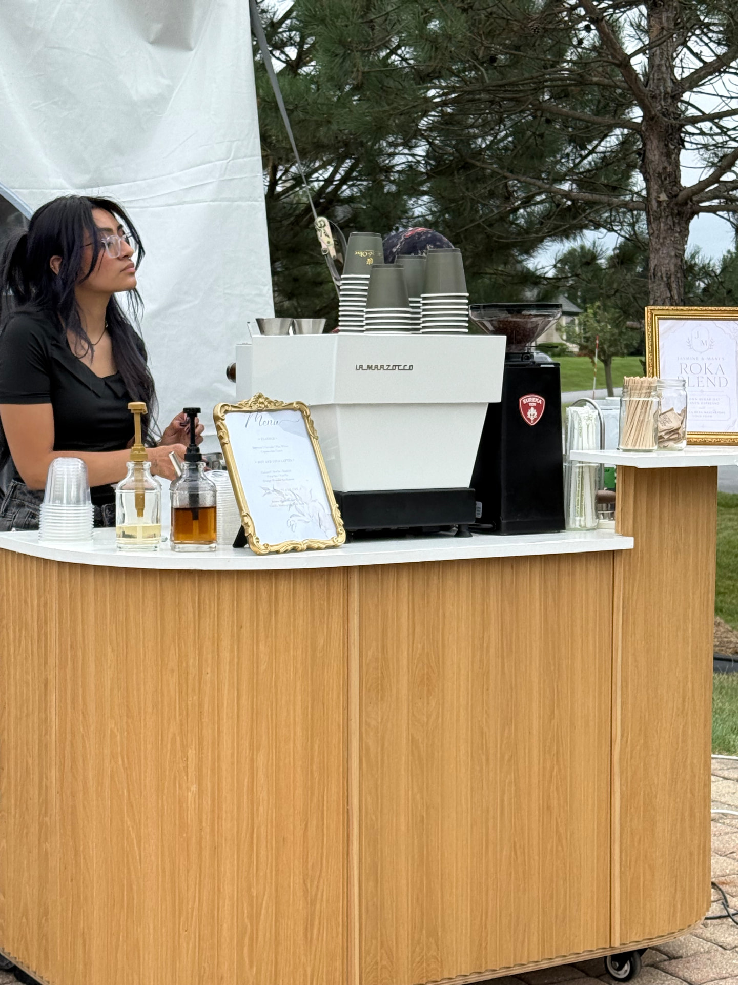 Barista serving guests from the Citrus & Olive Flutes cart at an engagement party, with a white La Marzocco espresso machine, olive-green branded cups and a framed couple's menu sign